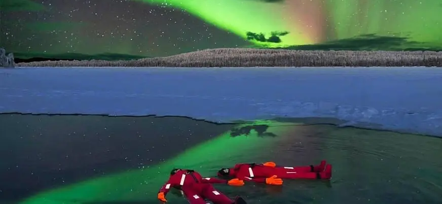 Dos personas con trajes térmicos rojos flotando en agua helada bajo una aurora boreal verde en un paisaje ártico, con nieve y bosque al fondo.