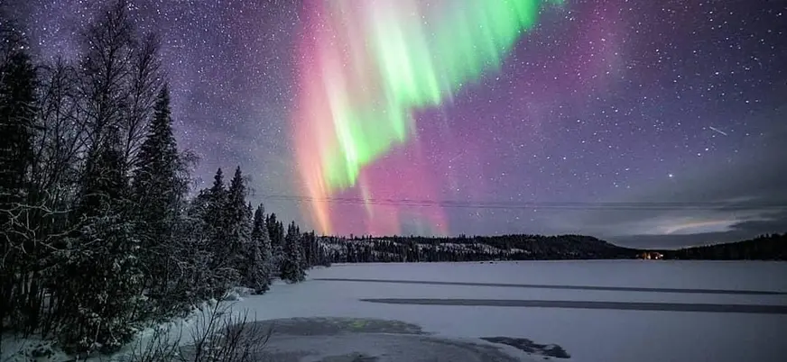 Aurora boreal con tonos verdes y rosados iluminando el cielo nocturno sobre un lago congelado y bosque nevado en un paisaje ártico.