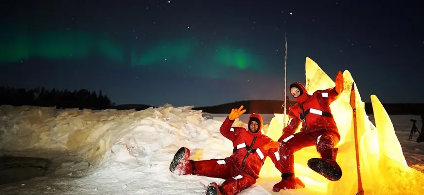 Dos personas con trajes térmicos rojos sentadas sobre nieve iluminada por luz cálida, bajo un cielo nocturno con aurora boreal verde en un paisaje ártico.