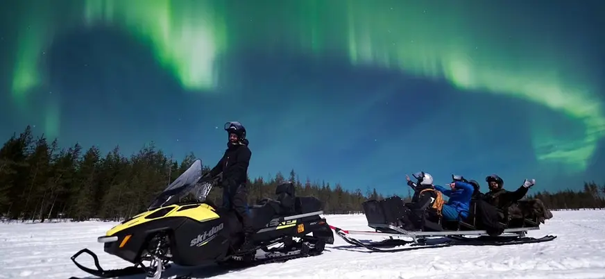 Motonieve sobre nieve con trineo y personas, bajo auroras boreales verdes en el cielo nocturno.