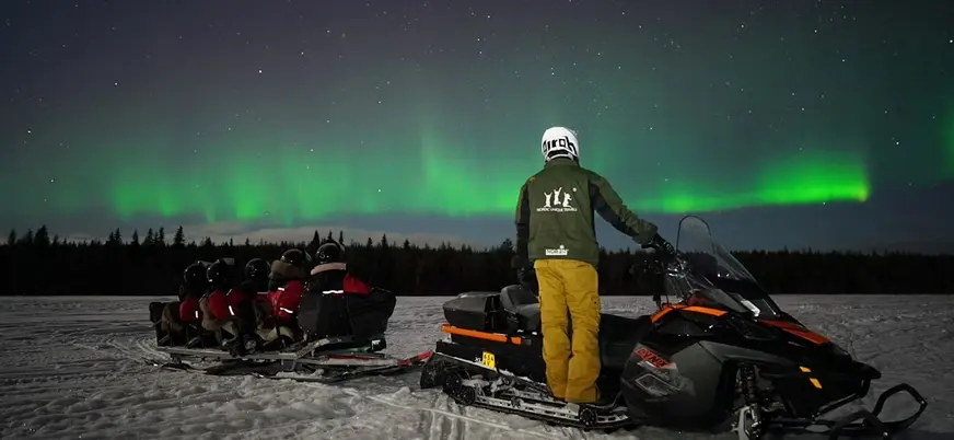 Motonieve negra y naranja con trineo sobre nieve, bajo auroras boreales verdes en el cielo nocturno.