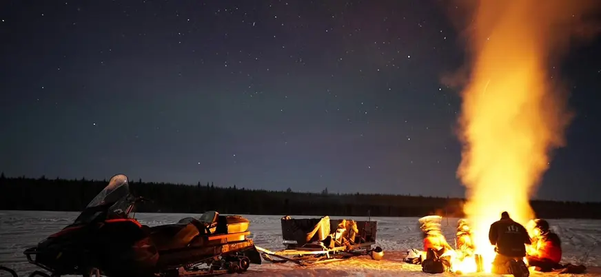 Grupo alrededor de una gran fogata en la nieve, junto a motonieve y trineos, bajo un cielo nocturno estrellado.