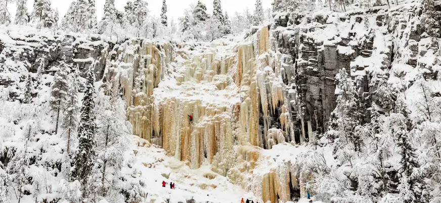 Gran cascada congelada con tonos amarillos y blancos, rodeada de bosque nevado, con varias personas en la base y un escalador en la pared de hielo.