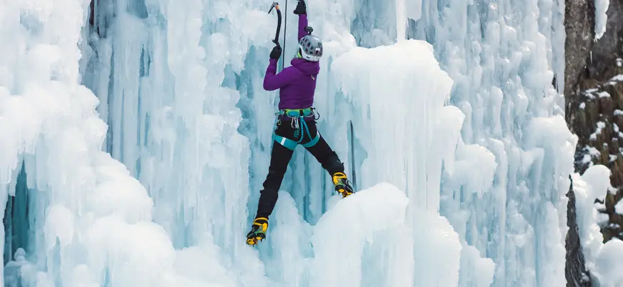 Persona escalando una pared de hielo con crampones, arnés y dos piolets, sobre una superficie congelada con formaciones irregulares.