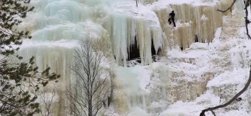 Persona escalando una gran cascada congelada con cuerdas, rodeada de nieve y árboles invernales.