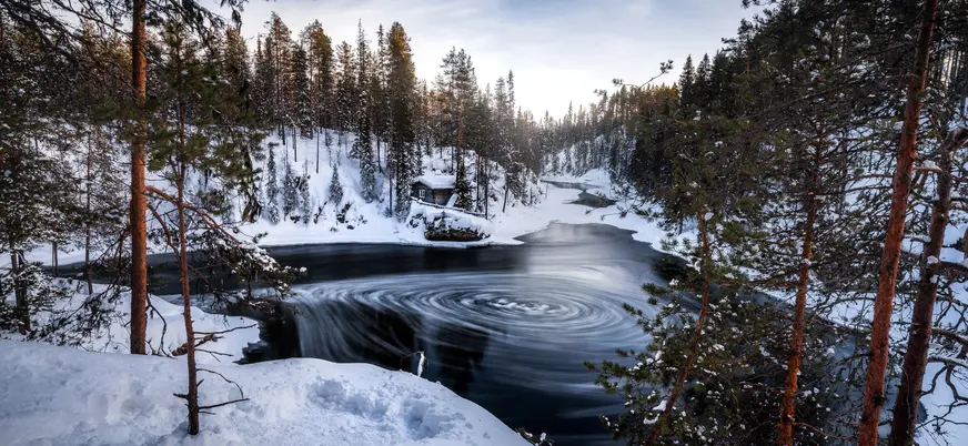 Río parcialmente congelado con remolinos en el agua, rodeado de bosque nevado y una cabaña de madera, bajo un cielo azul claro."