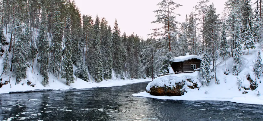 Cabaña de madera junto a un río parcialmente congelado, rodeada de bosque nevado bajo un cielo claro.