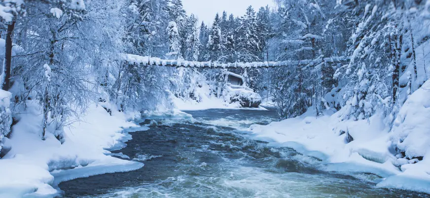 Río con corriente rodeado de bosque nevado, con un puente colgante cubierto de nieve y una cabaña al fondo.