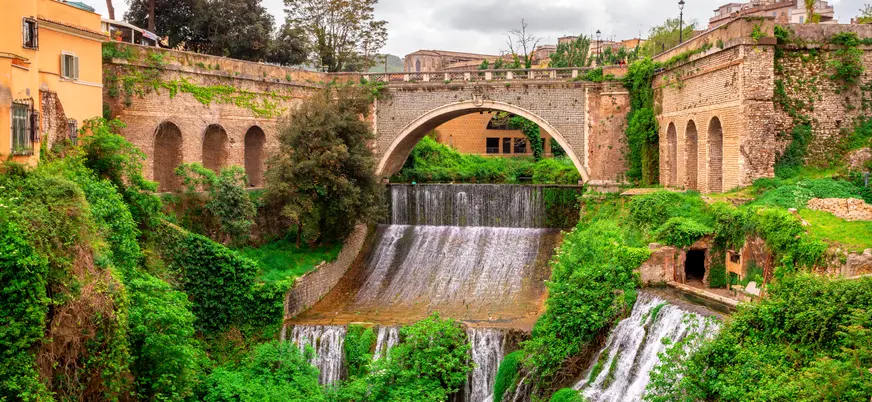 Cascada escalonada bajo puente histórico rodeado de vegetación y arquitectura tradicional.
