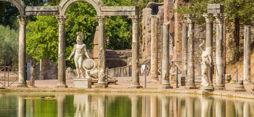 Estatuas y columnas romanas reflejadas en estanque rodeado de ruinas y vegetación en Tívoli.