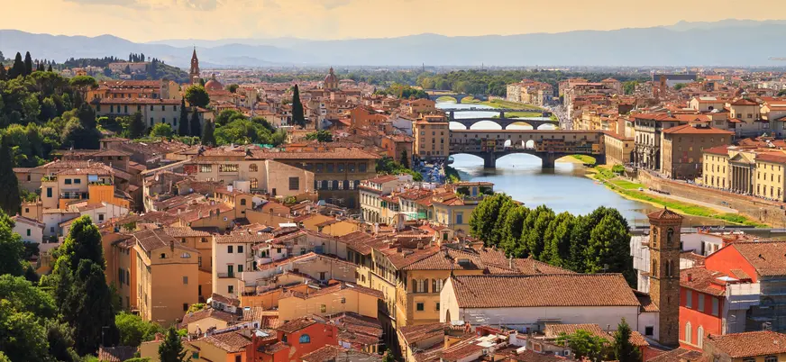 Vista panorámica de Florencia, Italia, mostrando el río Arno con varios puentes, incluido el famoso Ponte Vecchio, rodeado de edificios históricos y tejados rojizos bajo un cielo cálido al atardecer.