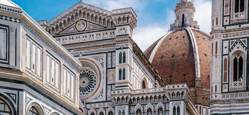 Detalle arquitectónico de la Catedral de Santa María del Fiore en Florencia, Italia, mostrando la fachada con mármol policromado y la cúpula de Brunelleschi bajo un cielo azul.