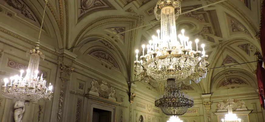 Salón elegante del Palacio Pitti en Florencia, con techos abovedados decorados, esculturas clásicas y grandes lámparas de araña de cristal iluminadas.