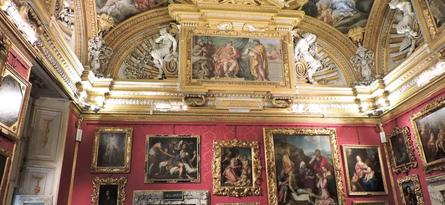 Sala del Palacio Pitti en Florencia con paredes rojas cubiertas de pinturas renacentistas en marcos dorados, techo decorado con frescos y molduras ornamentadas en oro.