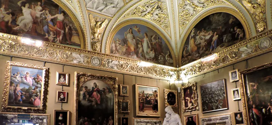 Interior del Palacio Pitti en Florencia, Italia: sala decorada con frescos barrocos en techos abovedados, molduras doradas, numerosas pinturas clásicas en marcos ornamentados y una escultura de mármol blanco.