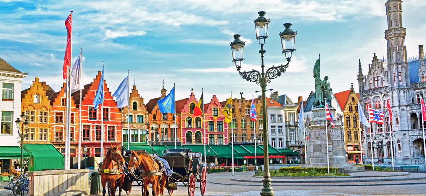 Plaza Mayor de Brujas con edificios flamencos de colores, banderas, farola antigua y carruaje tirado por caballos, bajo un cielo parcialmente nublado.