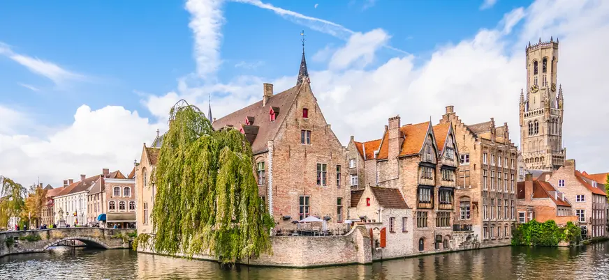 Canal de Brujas en Bélgica con edificios medievales de ladrillo y techos inclinados, terraza junto al agua y un árbol frondoso, bajo un cielo azul con nubes.