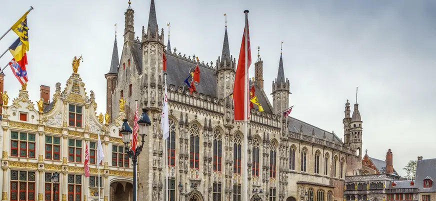 Plaza Burg en Brujas, Bélgica, con el Ayuntamiento gótico y edificios históricos adornados con banderas, bajo un cielo nublado.