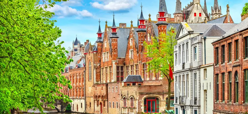 Canal de Brujas en Bélgica rodeado de edificios históricos de ladrillo, puentes y vegetación, con reflejos en el agua bajo un cielo parcialmente nublado.