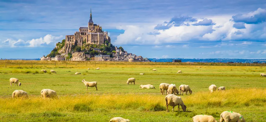 Mont Saint-Michel en Normandía, Francia, rodeado de praderas verdes con ovejas pastando bajo cielo parcialmente nublado.