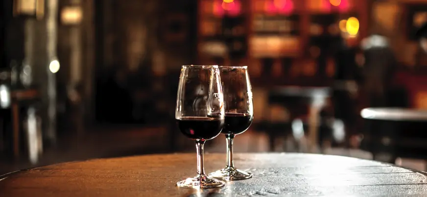 Dos copas de vino tinto sobre barrica en una bodega iluminada.