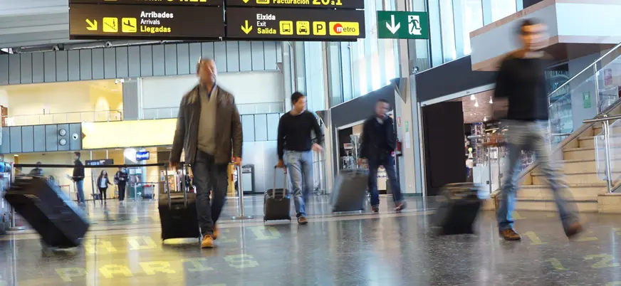 Interior de un aeropuerto con señalización de facturación y salidas, personas caminando con maletas de viaje.