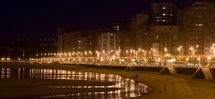 Vista nocturna del paseo marítimo y la playa de San Lorenzo en Gijón, con luces reflejadas sobre la arena mojada