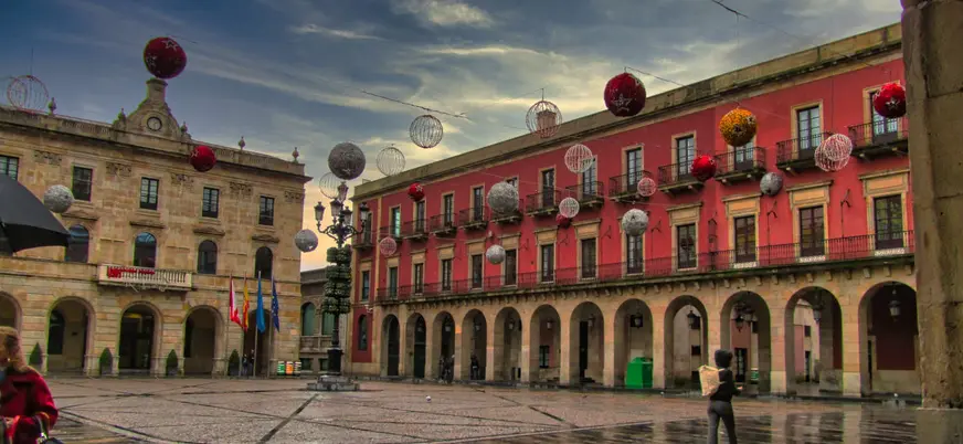 Plaza Mayor de Gijón con decoración navideña y edificios históricos, reflejados en el suelo mojado tras la lluvia