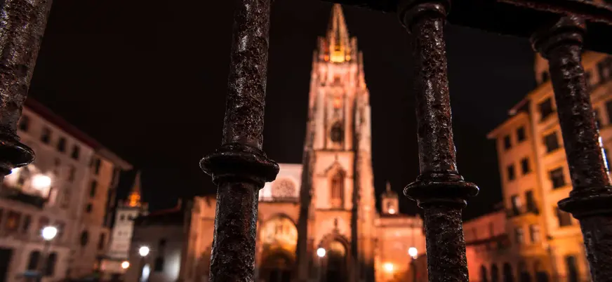 Vista nocturna de la Catedral de San Salvador de Oviedo a través de una barandilla, con la torre gótica iluminada en el centro.
