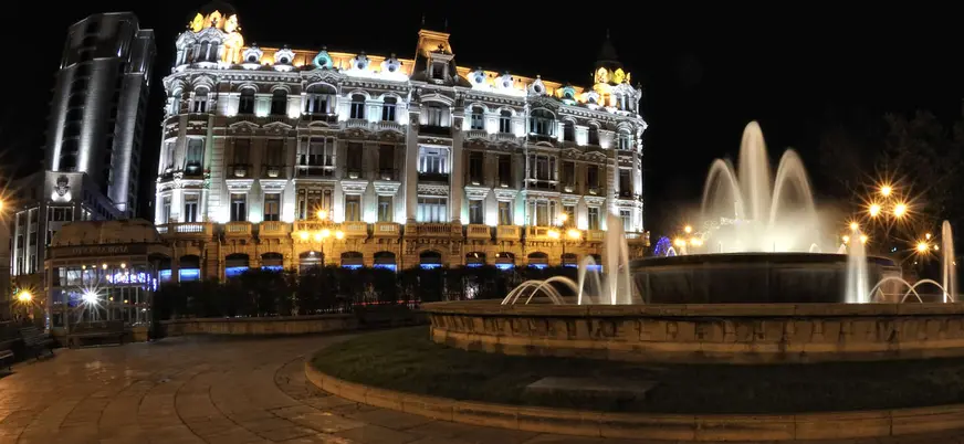 Fuente iluminada de la Plaza de la Escandalera en Oviedo frente al edificio histórico del Banco Herrero, una vista nocturna emblemática.