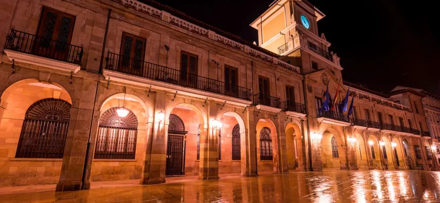 Ayuntamiento de Oviedo iluminado de noche con reflejos en el suelo mojado.