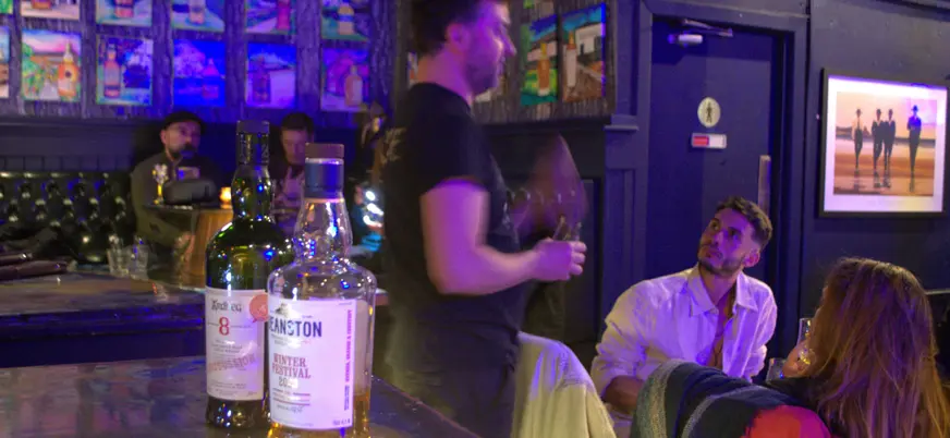 Sesión de cata de whisky escocés en un bar, con dos botellas de whisky en primer plano y personas participando en la actividad.