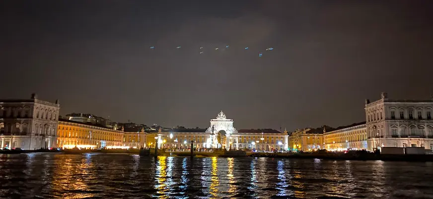 Vista nocturna de la Plaza del Comercio en Lisboa iluminada, con reflejos de luces en el río Tajo y cielo oscuro.