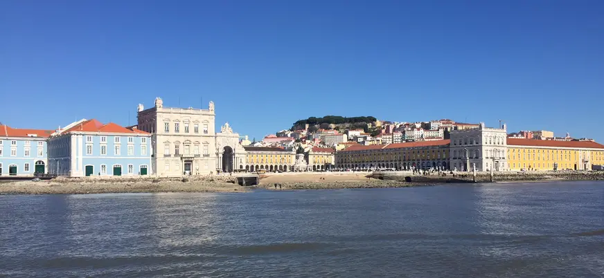 Vista panorámica de la Plaza del Comercio en Lisboa desde el río Tajo, con edificios históricos de fachadas amarillas y azules, colinas al fondo y cielo despejado.