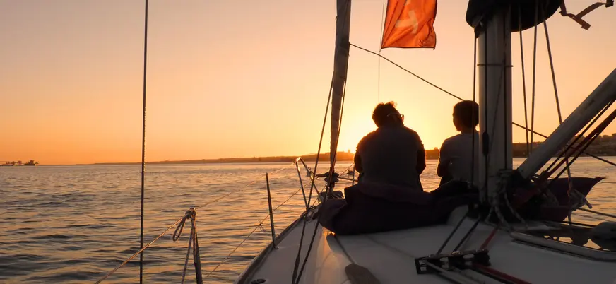 Dos personas sentadas en la proa de un velero durante la puesta de sol sobre el río Tajo en Lisboa, con el cielo iluminado en tonos cálidos.