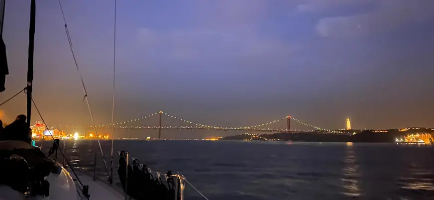 Vista nocturna del Puente 25 de Abril en Lisboa iluminado, con reflejos de luces en el río Tajo bajo un cielo oscuro.