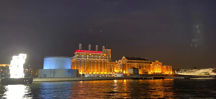 Vista nocturna de edificios históricos iluminados en la ribera del río Tajo en Lisboa, con reflejos de luces en el agua y cielo oscuro.
