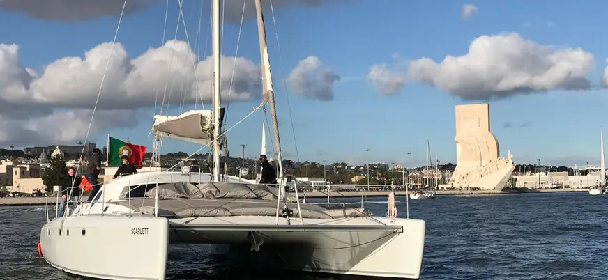Catamarán navegando por el río Tajo en Lisboa con bandera de Portugal, cielo parcialmente nublado y el Monumento a los Descubrimientos visible al fondo.