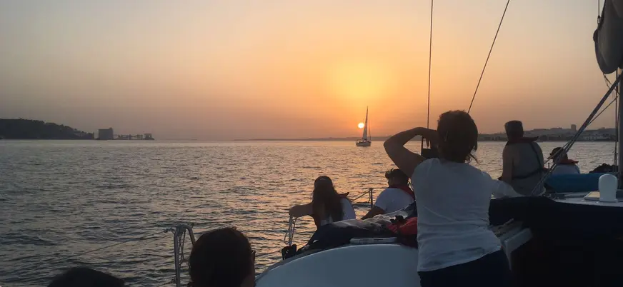 Grupo de personas disfrutando de un paseo en velero durante la puesta de sol sobre el río Tajo en Lisboa, con el cielo iluminado en tonos cálidos y reflejos en el agua.