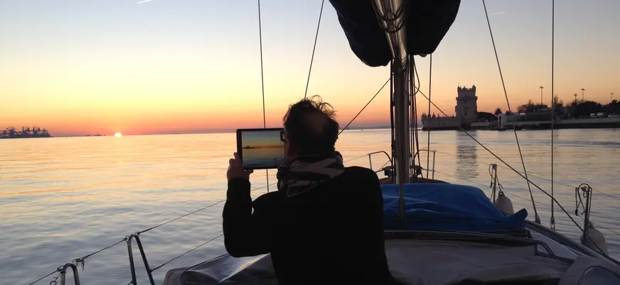 Persona sentada en la cubierta de un velero durante la puesta de sol sobre el río Tajo en Lisboa, con el cielo iluminado en tonos cálidos.
