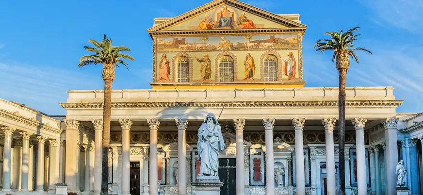 Fachada de la Basílica de San Pedro Extramuros en Roma, con columnas clásicas, mosaicos en el frontón y jardines con palmeras bajo cielo despejado.