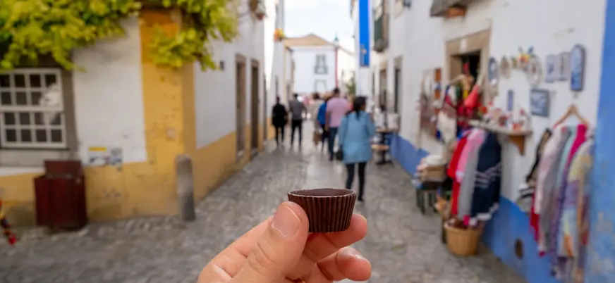 Mano sosteniendo un vaso de chocolate con ginja en una calle típica de Óbidos.