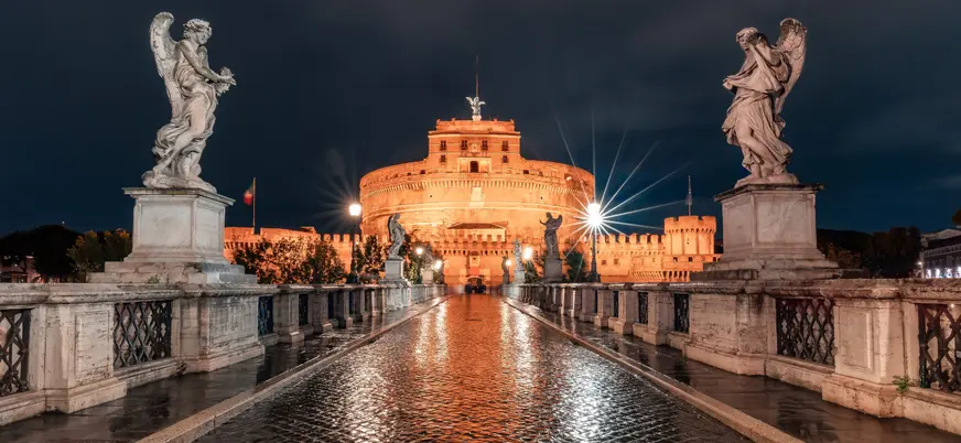 Castillo Sant'Angelo en Roma iluminado de noche, visto desde el Puente Sant'Angelo con estatuas barrocas y reflejos en el pavimento mojado.