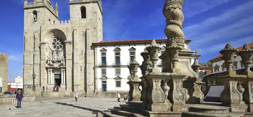 Fachada de la Catedral de Oporto y su plaza con columna barroca bajo un cielo azul.