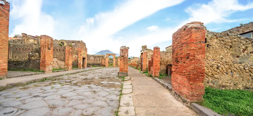 Calle romana empedrada en Pompeya con columnas y el Vesubio al fondo.
