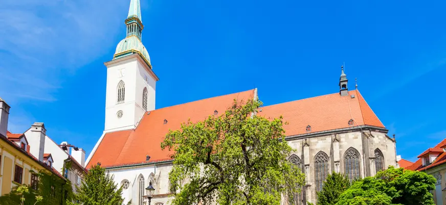 Catedral de San Martín en Bratislava, con su torre de aguja verde y tejado rojo, rodeada de árboles bajo un cielo azul claro.