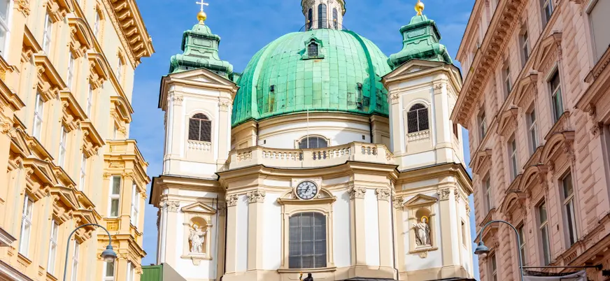 Fachada de la Iglesia de San Pedro en Viena, con su gran cúpula verde y torres barrocas iluminadas por el sol.