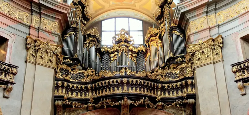 Órgano barroco dorado de la Iglesia de San Pedro en Viena, con tubos metálicos y elaboradas decoraciones talladas bajo una bóveda pintada.