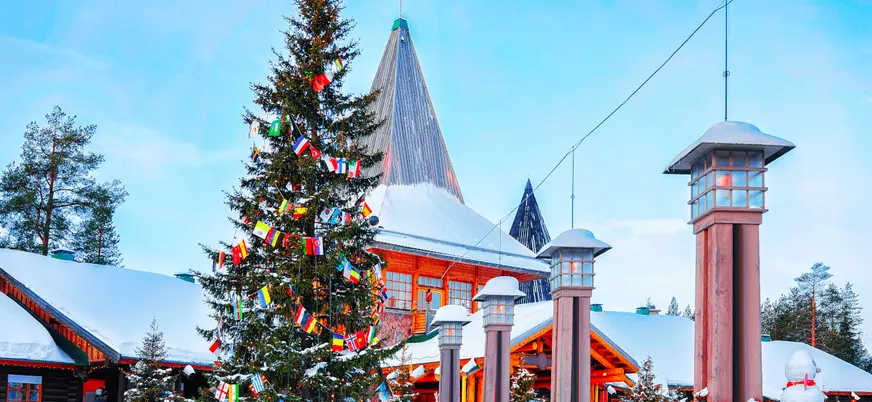 Árbol de Navidad decorado con banderas internacionales frente a una construcción de madera con techo nevado en la aldea de Santa Claus, Laponia, Finlandia.