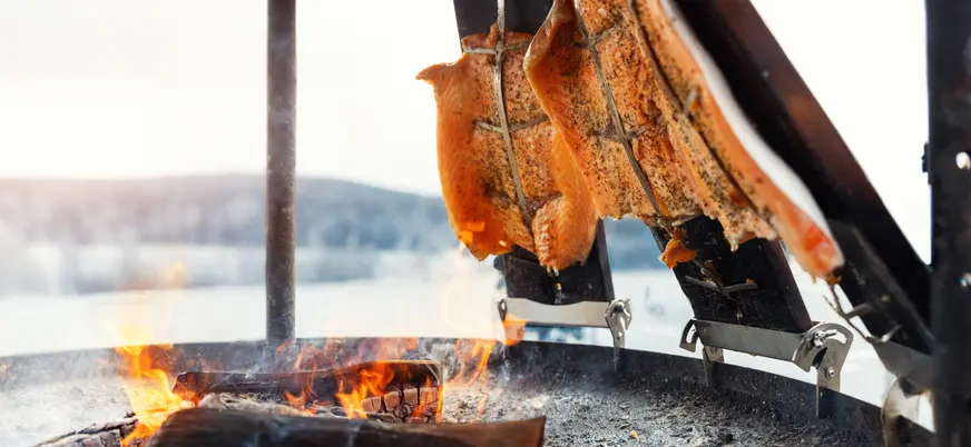Filetes de salmón sazonados asándose al fuego en una parrilla tradicional al aire libre, con brasas encendidas y paisaje invernal de fondo.